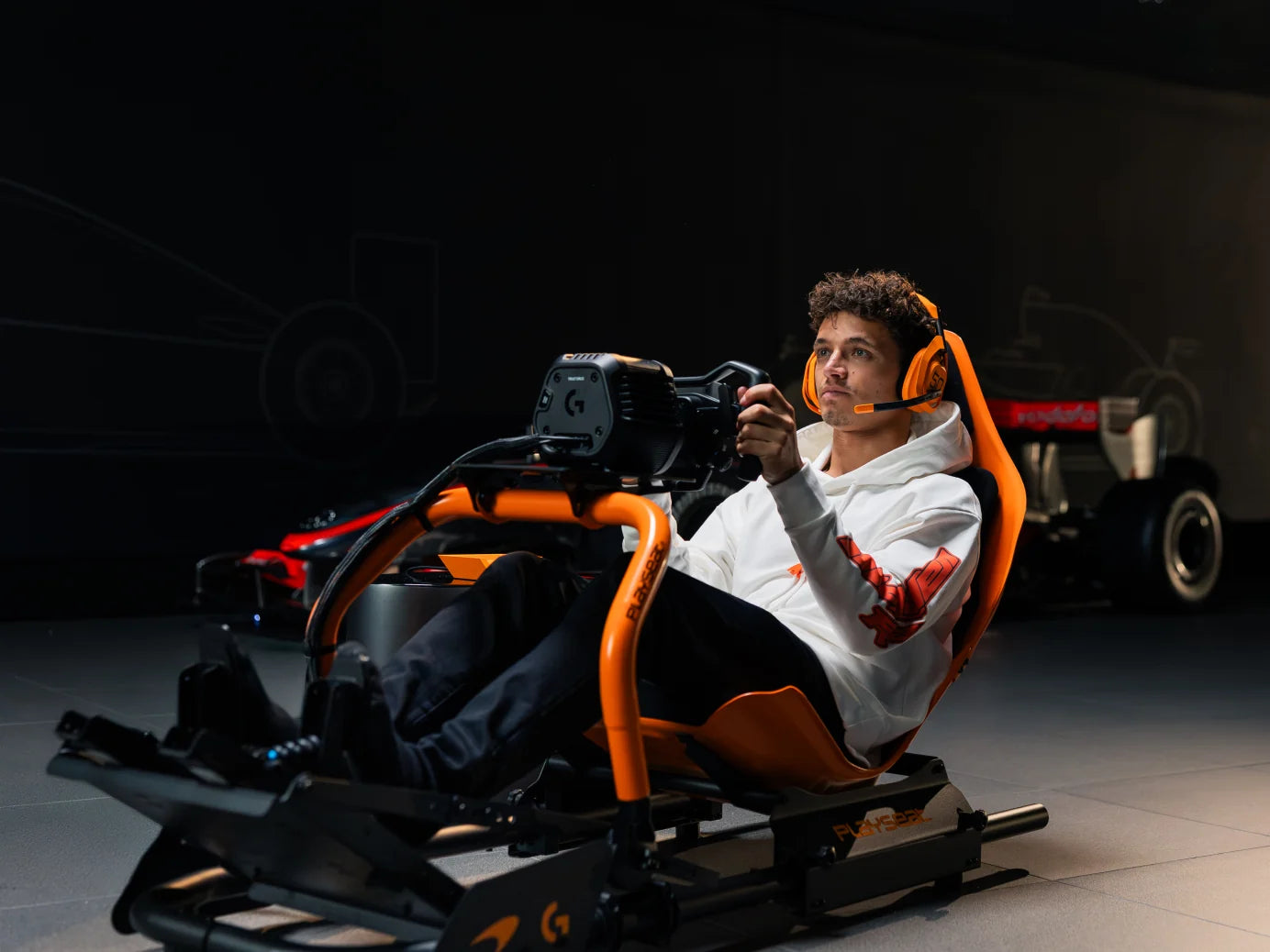 Driver racing in the Playseat Formula Instinct McLaren Racing Edition beside McLaren show cars